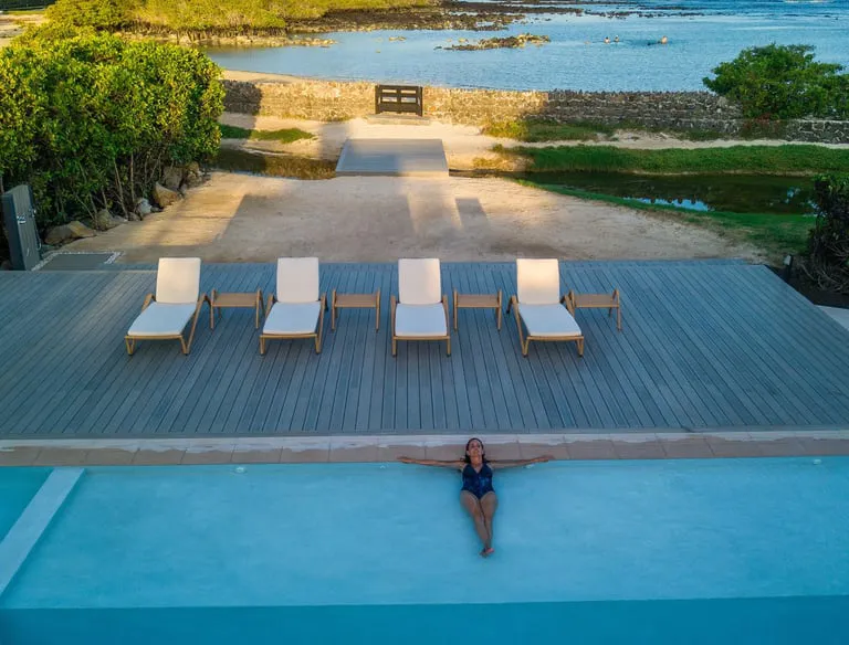 Person in a swimsuit sits at the pool edge with arms outstretched on a blue pool, wooden deck