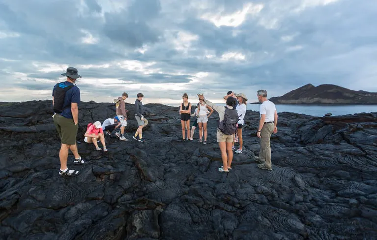 Tour group standing on glossy black lava rock by the coast, with clouds and distant hills overhead.