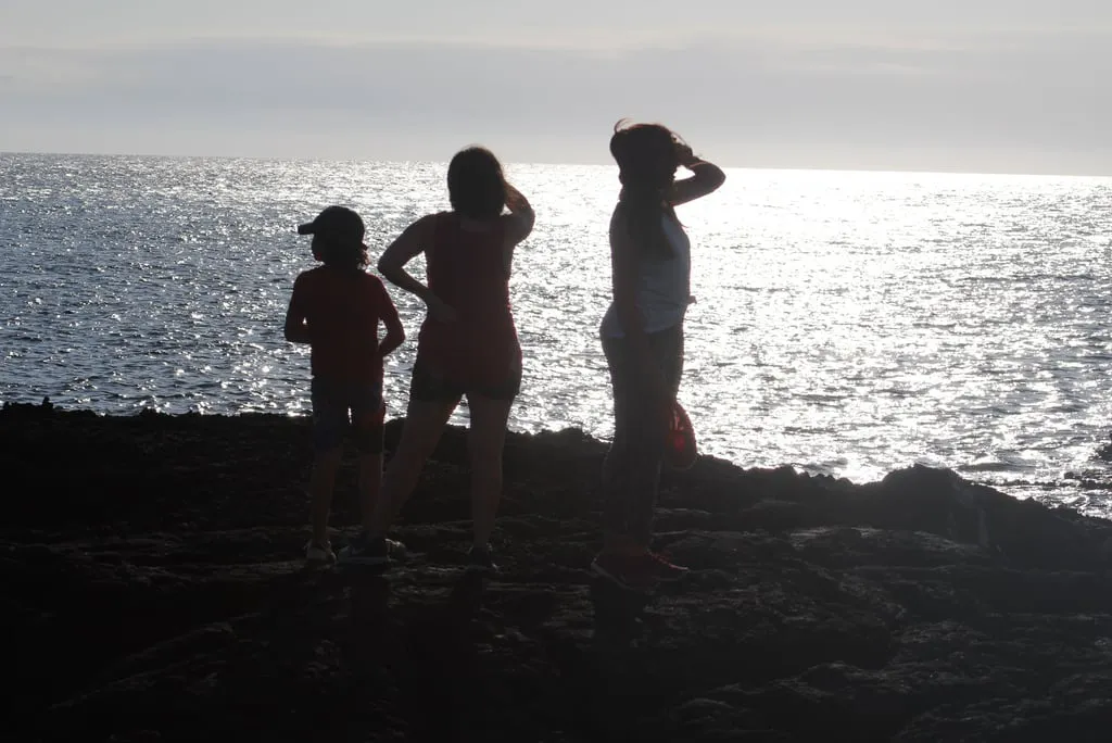 Silhouetted trio standing on a rocky beach, facing a shimmering sea at sunset.