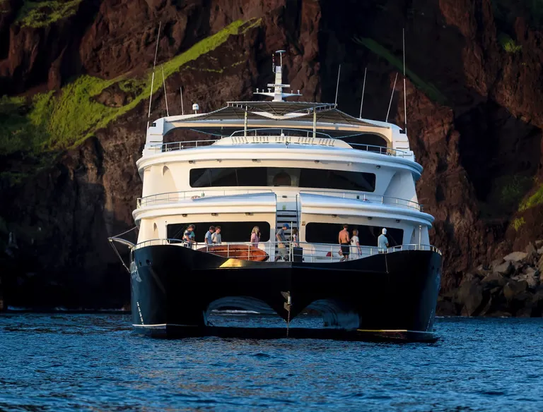 Large luxury motor yacht anchored near a rocky cliff, with people on the rear deck.