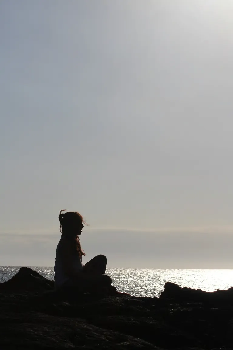 Silhouette of a person sitting cross-legged on rocky shore as the sun reflects on the ocean at sunset.