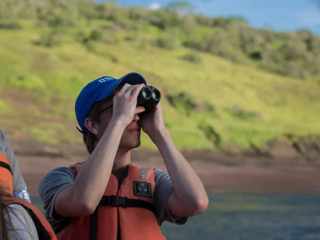 Person in an orange life jacket and blue cap uses binoculars while standing on a boat