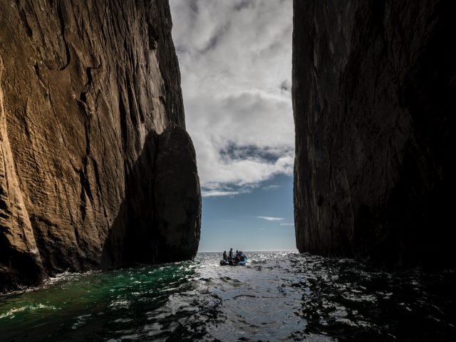 People in a small inflatable boat navigate a narrow sea inlet between towering rock cliffs