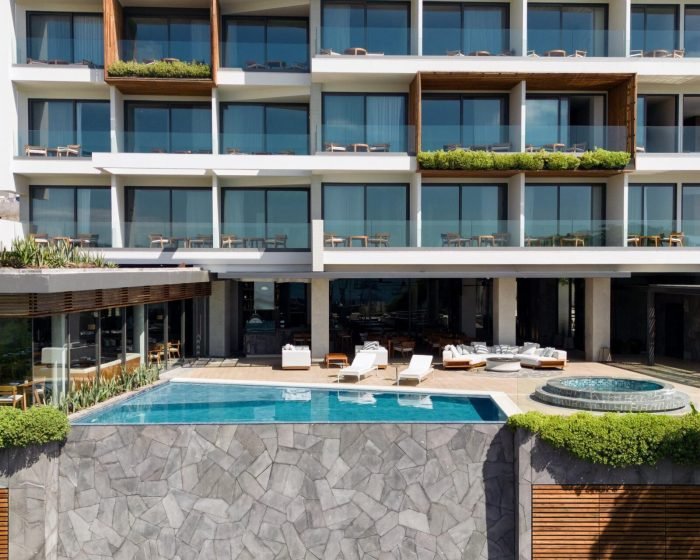 Modern multi-story building with glass balconies, overlooking a resort-style pool and terrace lounge areas on each level.