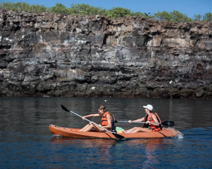 Two people in orange life jackets kayak together on calm water with a rocky cliff behind them.
